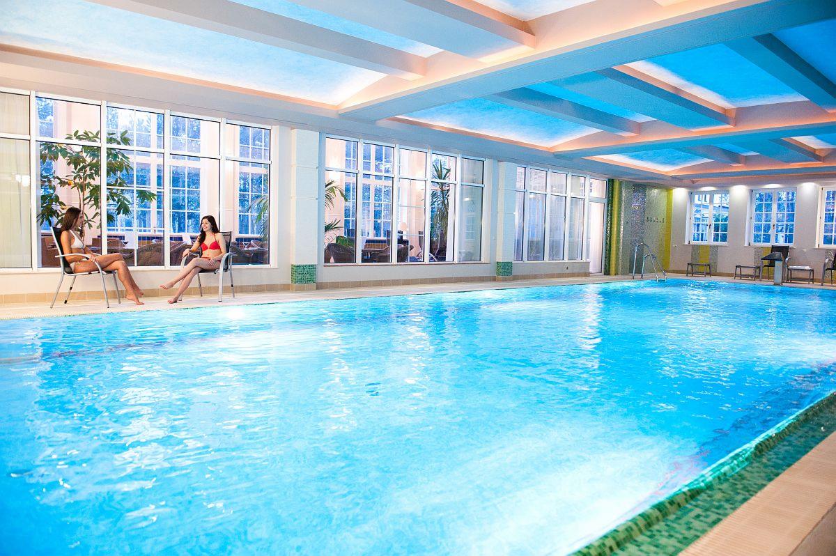 Jasny, przestronny basen kryty z dwoma kobietami siedzącymi na krzesłach przy oknie, relaksującymi się w bikini, duże okna z widokiem na zewnątrz, podświetlany sufit.