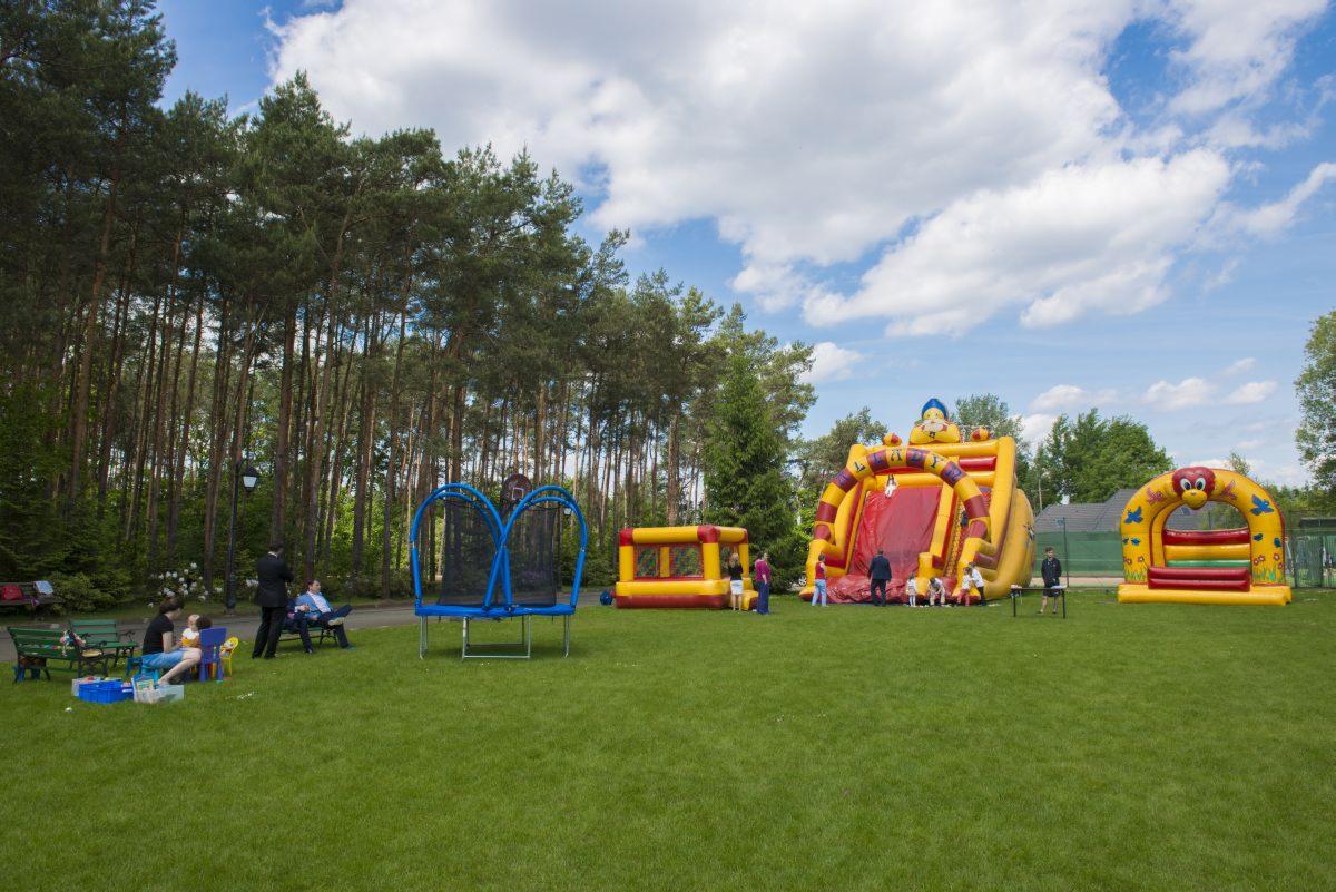 Rozległy plac zabaw na trawie z dmuchanym zamkiem ze zjeżdżalnią, tunelem z motywem motyla, trampoliną z podwójnym koszem i rodziną siedzącą na ławce w tle drzew.