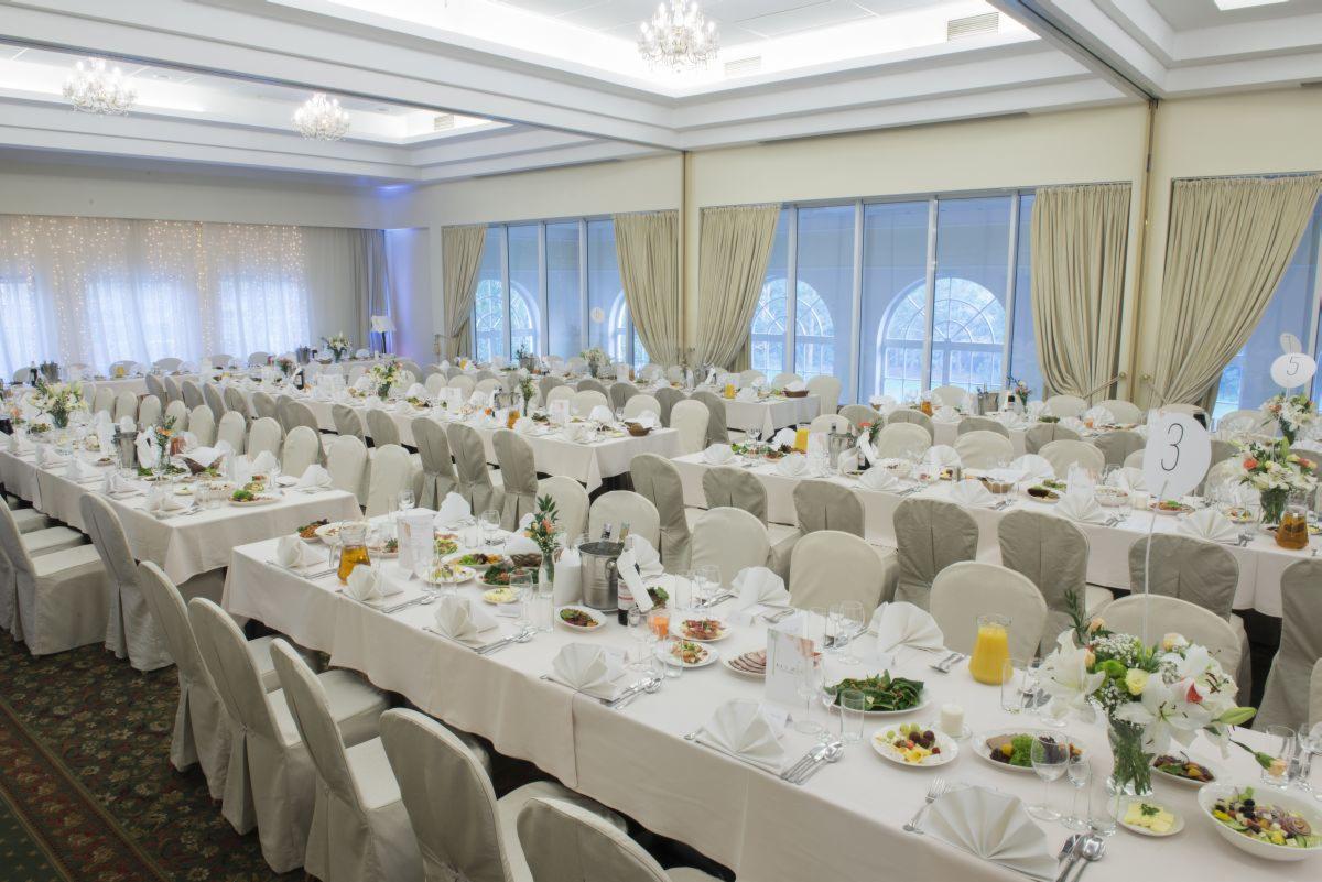 Uroczysta sala bankietowa z długimi stołami nakrytymi białymi obrusami, eleganckimi krzesłami, zastawą stołową, kwiatami i numeracją stołów, przygotowana na przyjęcie weselne lub inną imprezę...