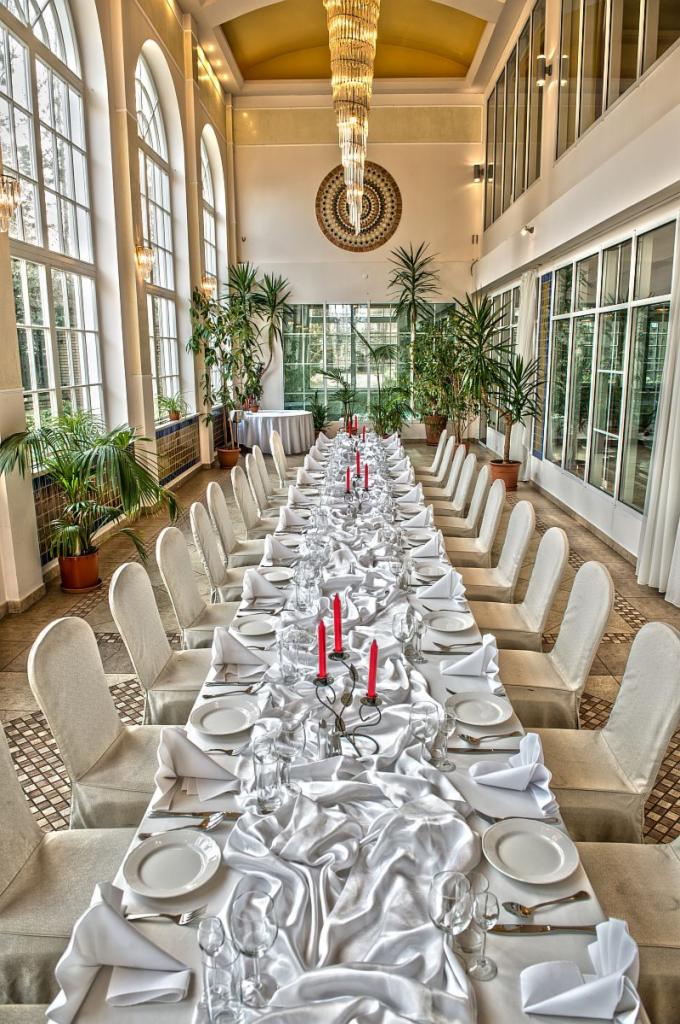 Długi stół bankietowy nakryty białym obrusem i zastawą, krzesła z białymi pokrowcami, czerwone świece w srebrnych świecznikach, wysokie okna w tle, kryształowy żyrandol i rośliny doniczkowe...