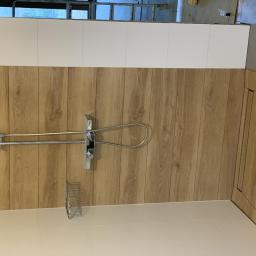 MAGGROUP Sp. z o.o. - Ukończona łazienka z panelami imitującymi drewno, białymi płytkami powyżej, chromowaną baterią prysznicową z deszczownicą i półką na kosmetyki, widoczne niedokończone prace na górze.