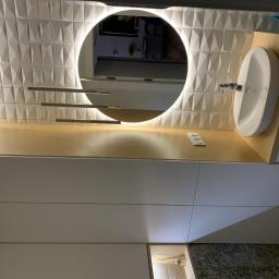 MAGGROUP Sp. z o.o. - Nowoczesna łazienka z owalną umywalką nablatową, podświetlanym okrągłym lustrem i trójwymiarową białą ścianą. Widoczne minimalistyczne oświetlenie i drewniany blat pod umywalką.