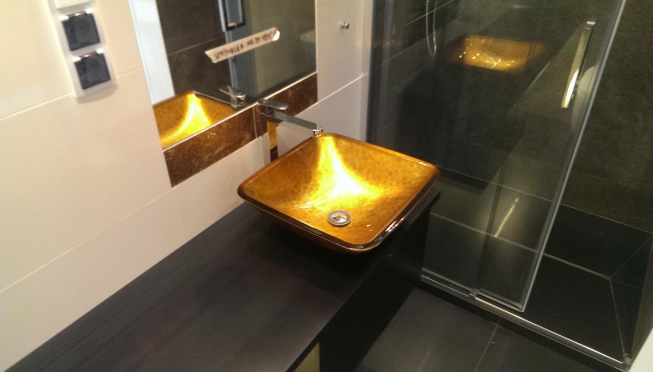 Umywalka w kolorze złota, kwadratowa, z chromowaną baterią, zamontowana na czarnym blacie w łazience z kabiną prysznicową w tle.