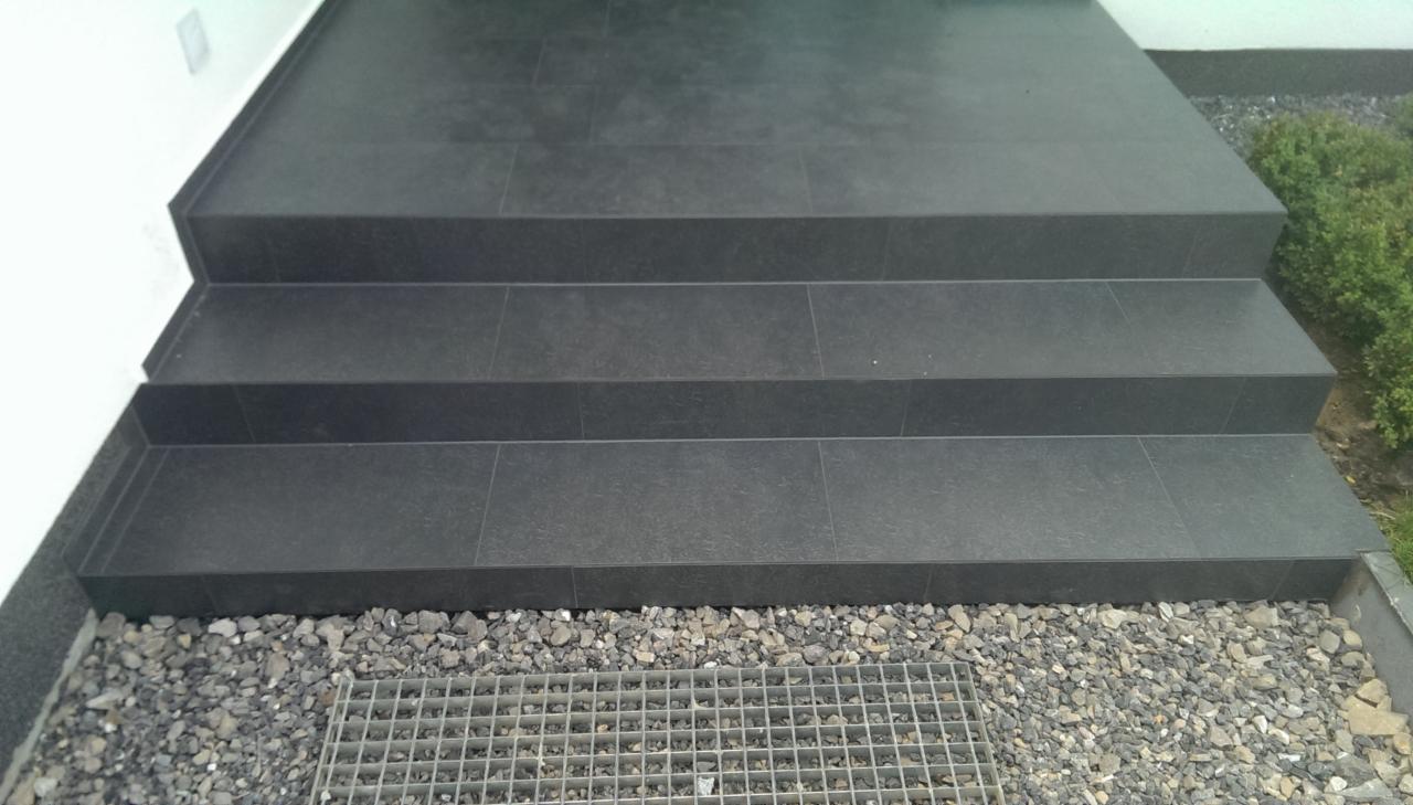 Zewnętrzne schody wejściowe wyłożone ciemnoszarą, matową płytką, z widoczną kratką odpływową i drobnym żwirem.