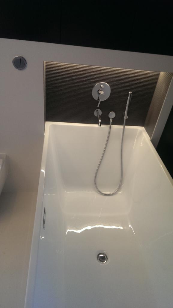 Biała, prostokątna wanna z chromowaną armaturą, w tym bateria i słuchawka prysznicowa, zamontowana na ścianie z ciemnoszarym panelem dekoracyjnym i subtelnym oświetleniem.