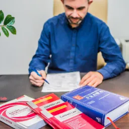 Mężczyzna w niebieskiej koszuli siedzi przy biurku, podpisując dokument. Na biurku leżą książki prawnicze w języku angielskim, w tym 'English Legal System', 'Company Law' i 'Encyclopedia of Law'.