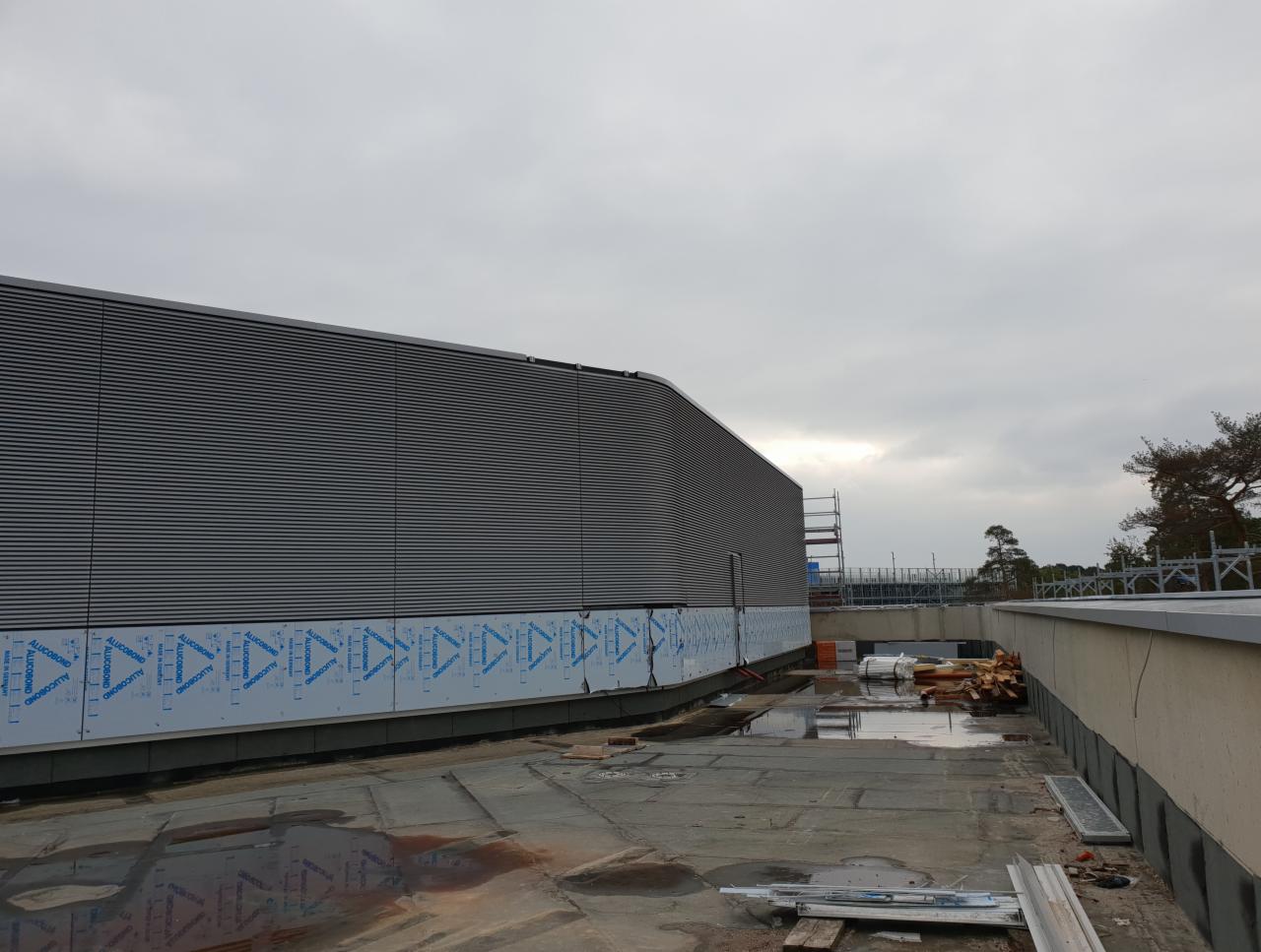 Montaż elewacji budynku przemysłowego z szarych paneli, widoczne fragmenty folii ochronnej na panelach przy ziemi, w tle rusztowanie i pochmurne niebo.