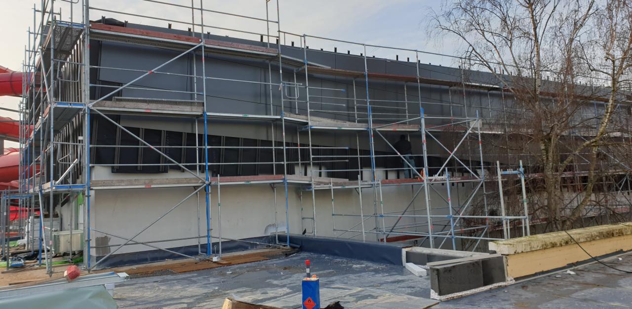 Budynek w trakcie ocieplania z widocznymi rusztowaniami, fragmentami elewacji w kolorze szarym i czarną izolacją termiczną, z boku widoczny fragment czerwonej zjeżdżalni wodnej.