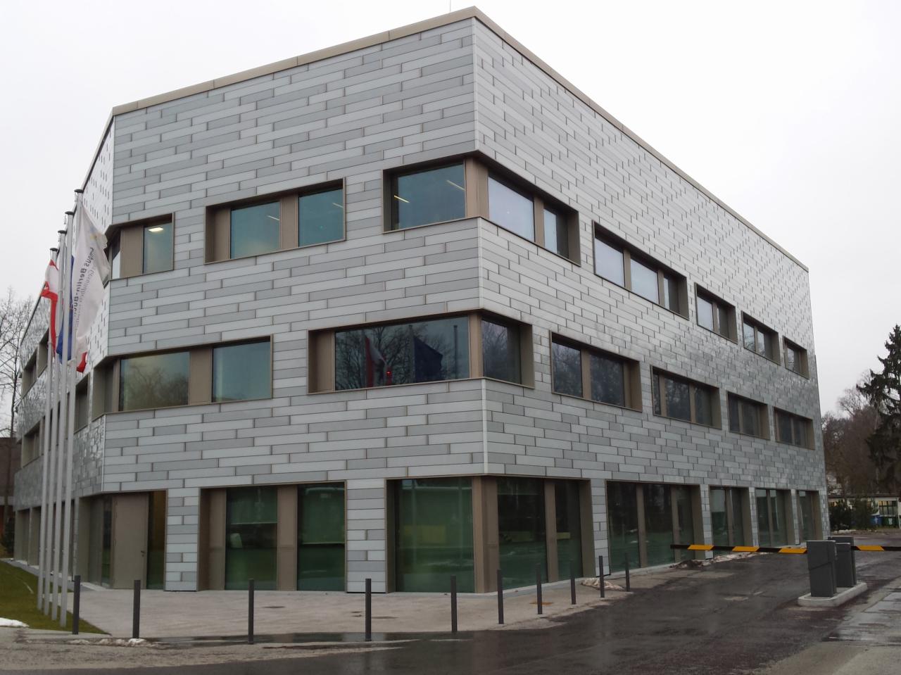 Nowoczesny budynek biurowy z szarą elewacją z płytek imitujących cegłę, widoczne okna z brązowymi ramami i maszty z flagami przed budynkiem, pochmurne niebo.