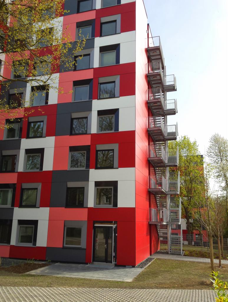 Wielopiętrowy budynek z nowoczesną elewacją w kratkę, w odcieniach czerwieni, szarości i bieli, z zewnętrzną klatką schodową i zielenią w tle.