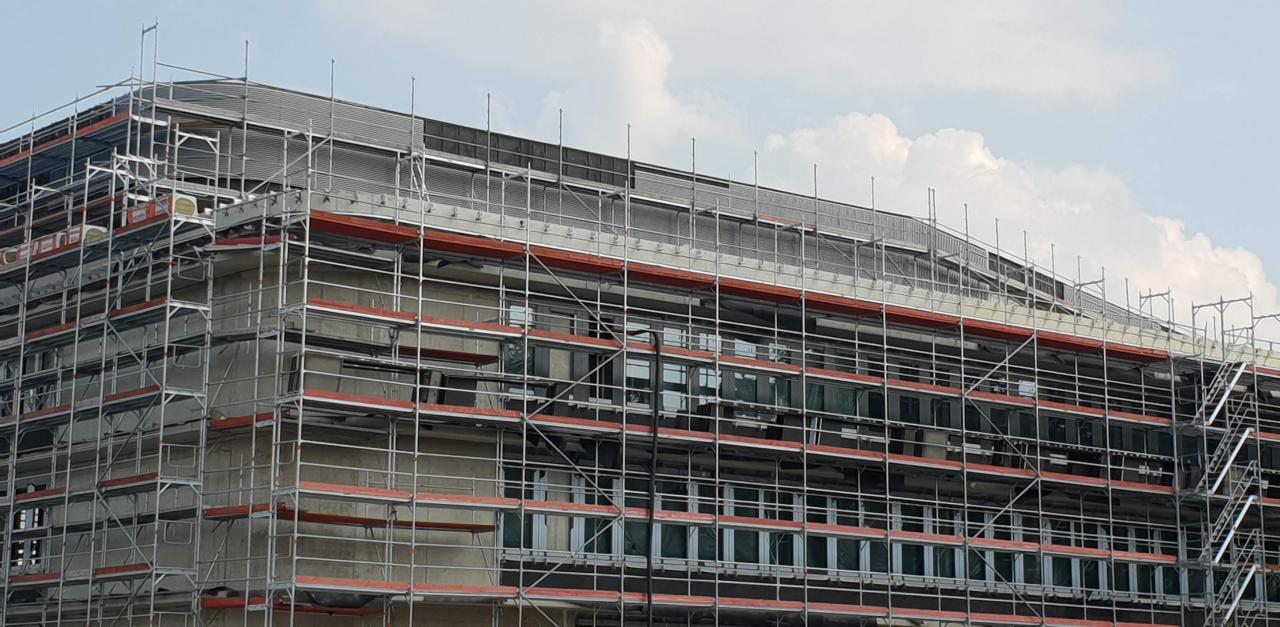 Budynek biurowy w trakcie prac elewacyjnych, osłonięty rusztowaniami z czerwonymi barierkami i widocznymi oknami, na tle jasnego nieba z chmurami.