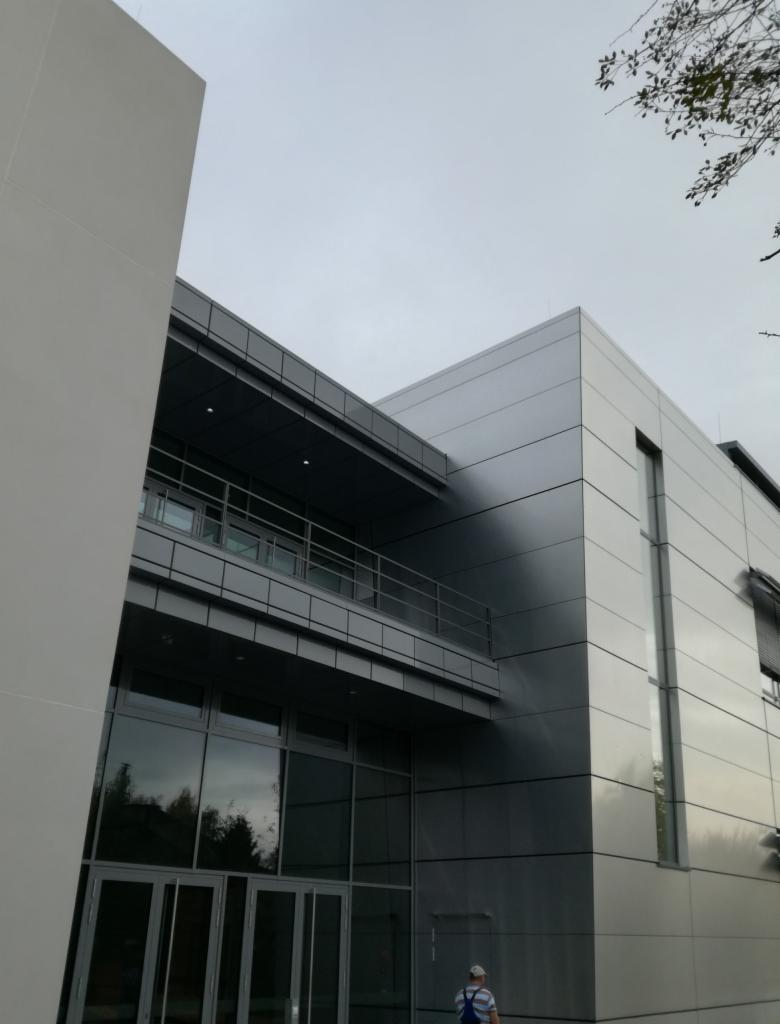 Nowoczesny budynek biurowy z elewacją z paneli metalowych, widoczne duże okna i balkon, fragment pracownika w dolnym rogu.