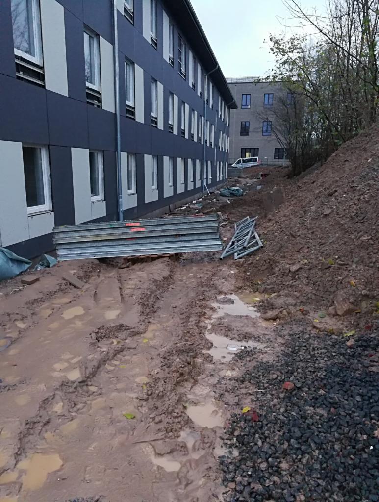 Budynek wielopiętrowy w trakcie prac elewacyjnych, widoczne rusztowania, błotniste podłoże i pryzma ziemi.