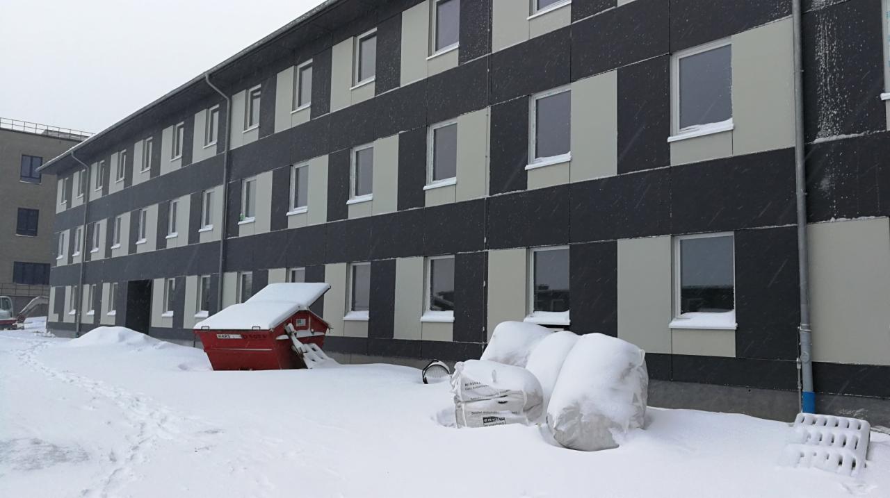 Budynek wielorodzinny z elewacją w odcieniach szarości, częściowo ocieplony, z workami materiałów budowlanych i kontenerem na gruz przykrytymi śniegiem podczas opadów śniegu.