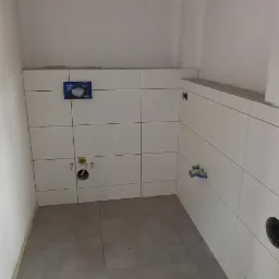 Łazienka w trakcie wykańczania: ułożone białe płytki na ścianach, widoczne instalacje wodne i kanalizacyjne, zamontowany stelaż podtynkowy WC, szare płytki podłogowe, surowa ściana z puszkami...