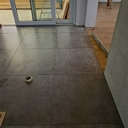 Probud - Układanie szarych, matowych płytek podłogowych w pomieszczeniu z przesuwnymi drzwiami balkonowymi, widoczna rolka taśmy malarskiej na podłodze i poziomica przy ścianie.