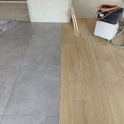 Probud - Połączenie szarych płytek podłogowych i paneli podłogowych w odcieniach drewna, widoczna maszyna do czyszczenia podłóg i surowy filar w tle, fragment remontowanego wnętrza.