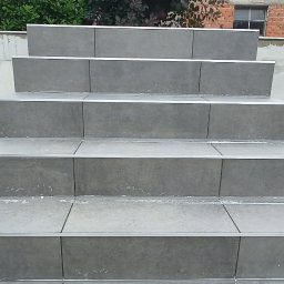 Probud - Schody zewnętrzne wyłożone szarą płytką z aluminiowymi listwami krawędziowymi, widoczne ślady zaprawy, tło stanowi betonowy mur i budynek z cegły.