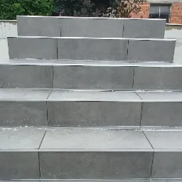 Schody zewnętrzne wyłożone szarą płytką z aluminiowymi listwami krawędziowymi, widoczne ślady zaprawy, tło stanowi betonowy mur i budynek z cegły.