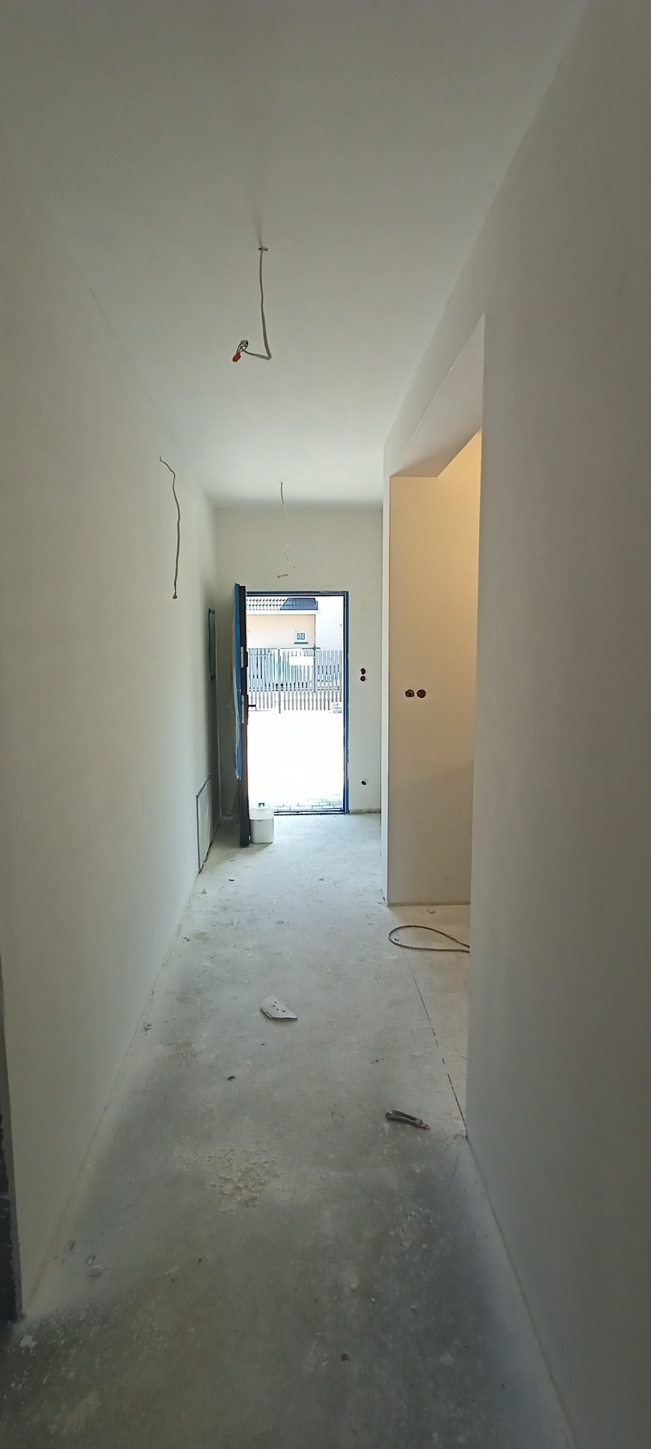 Korytarz w trakcie remontu: gładkie, pomalowane na biało ściany, surowa podłoga, wystające kable elektryczne, otwarte drzwi wejściowe z widokiem na zewnątrz.