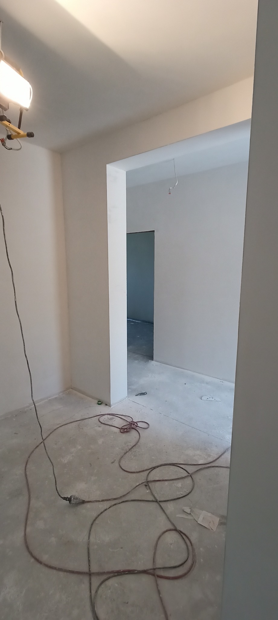 Wnętrze w trakcie malowania na biało, widoczny przewód elektryczny narzędzia na podłodze i lampa halogenowa oświetlająca ścianę.