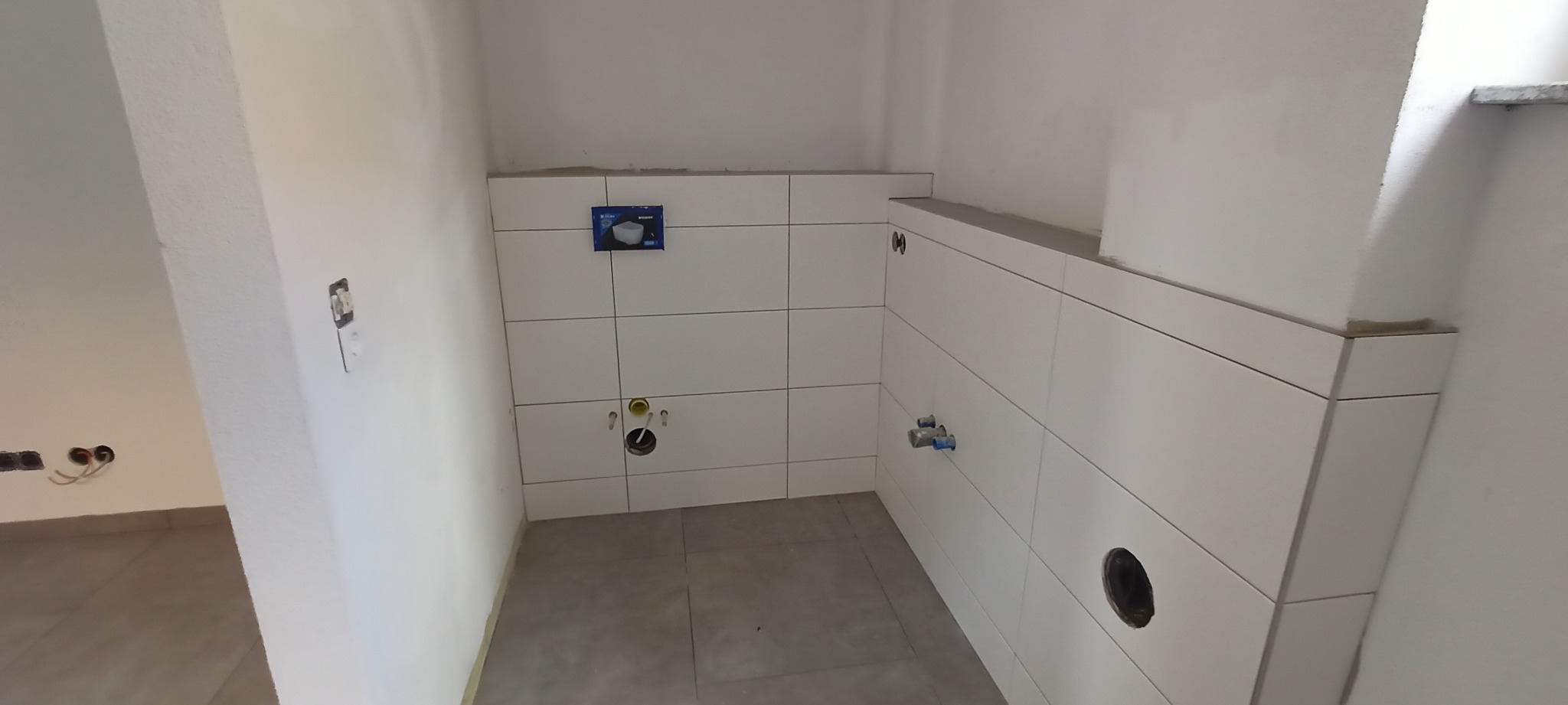 Łazienka w trakcie wykańczania: ułożone białe płytki na ścianach, widoczne instalacje wodne i kanalizacyjne, zamontowany stelaż podtynkowy WC, szare płytki podłogowe, surowa ściana z puszkami...