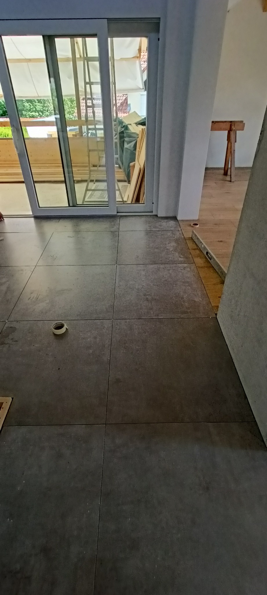 Układanie szarych, matowych płytek podłogowych w pomieszczeniu z przesuwnymi drzwiami balkonowymi, widoczna rolka taśmy malarskiej na podłodze i poziomica przy ścianie.