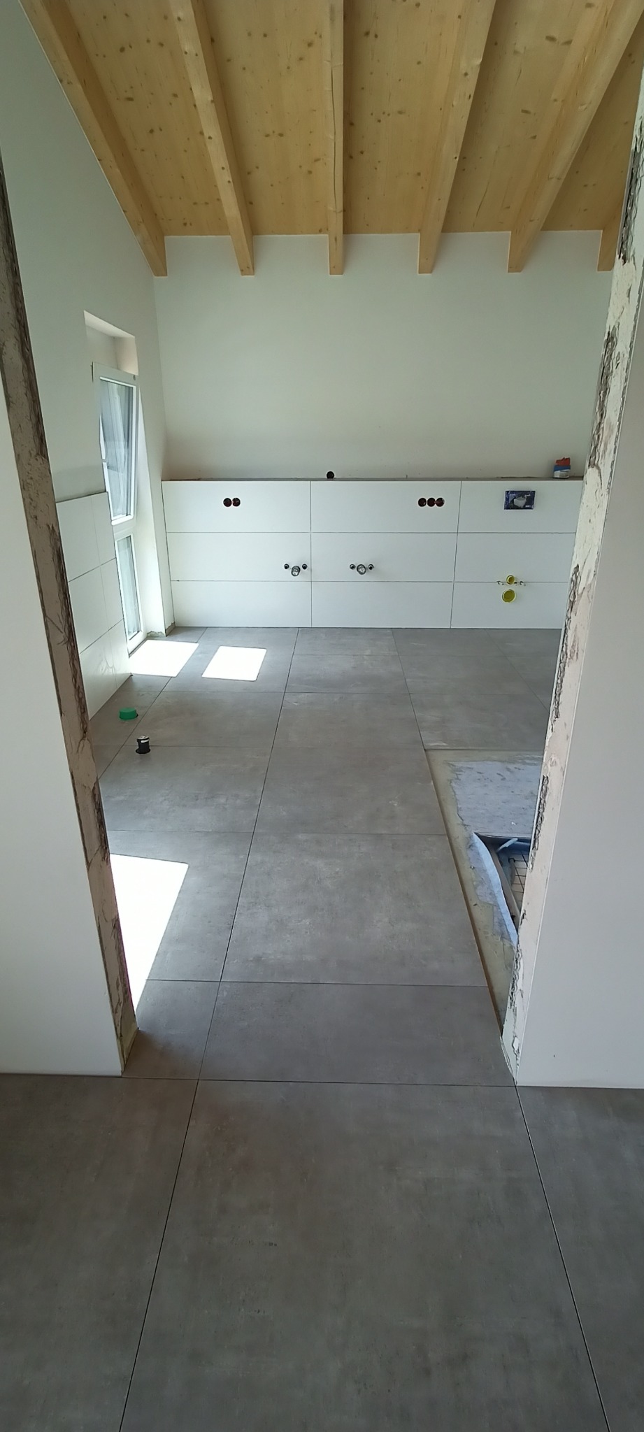 Szare płytki podłogowe ułożone w łazience w trakcie prac wykończeniowych, widoczne przygotowane punkty instalacyjne na ścianie i surowy drewniany strop.