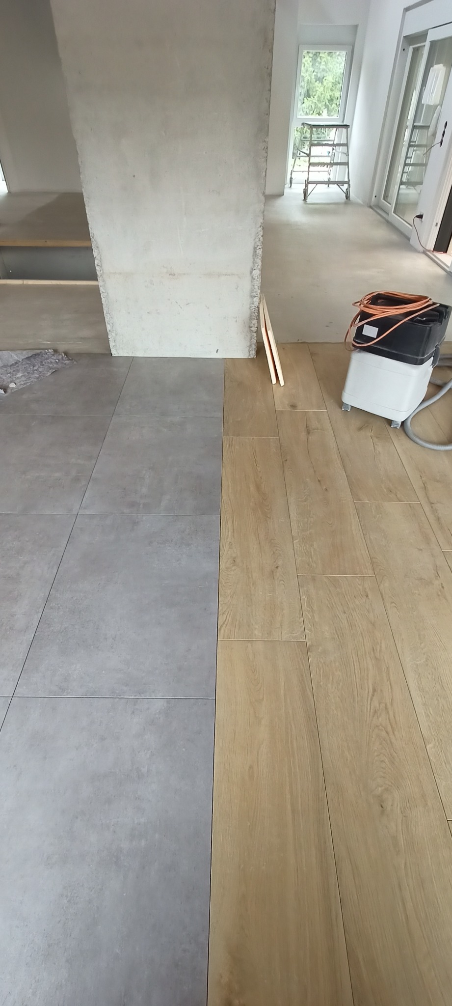 Połączenie szarych płytek podłogowych i paneli podłogowych w odcieniach drewna, widoczna maszyna do czyszczenia podłóg i surowy filar w tle, fragment remontowanego wnętrza.