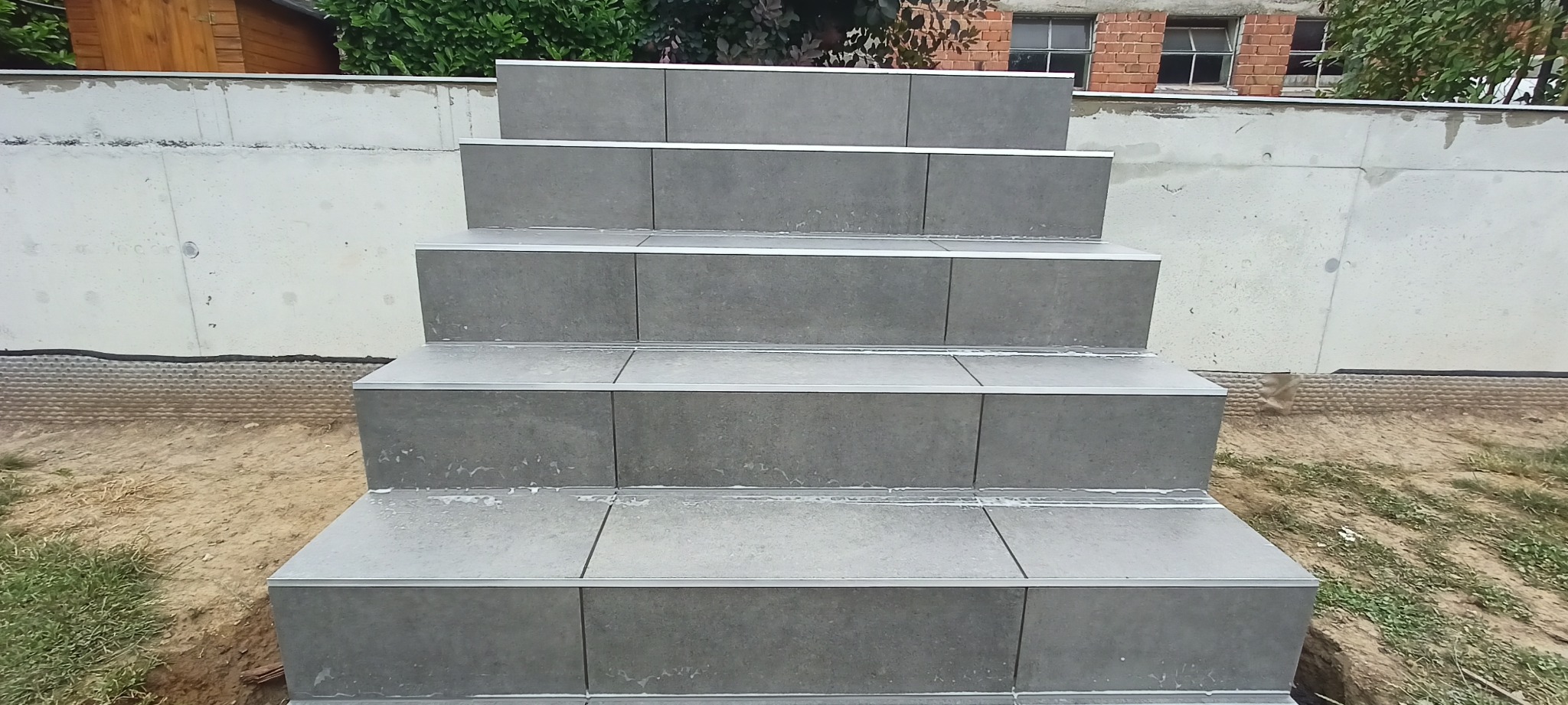 Schody zewnętrzne wyłożone szarą płytką z aluminiowymi listwami krawędziowymi, widoczne ślady zaprawy, tło stanowi betonowy mur i budynek z cegły.