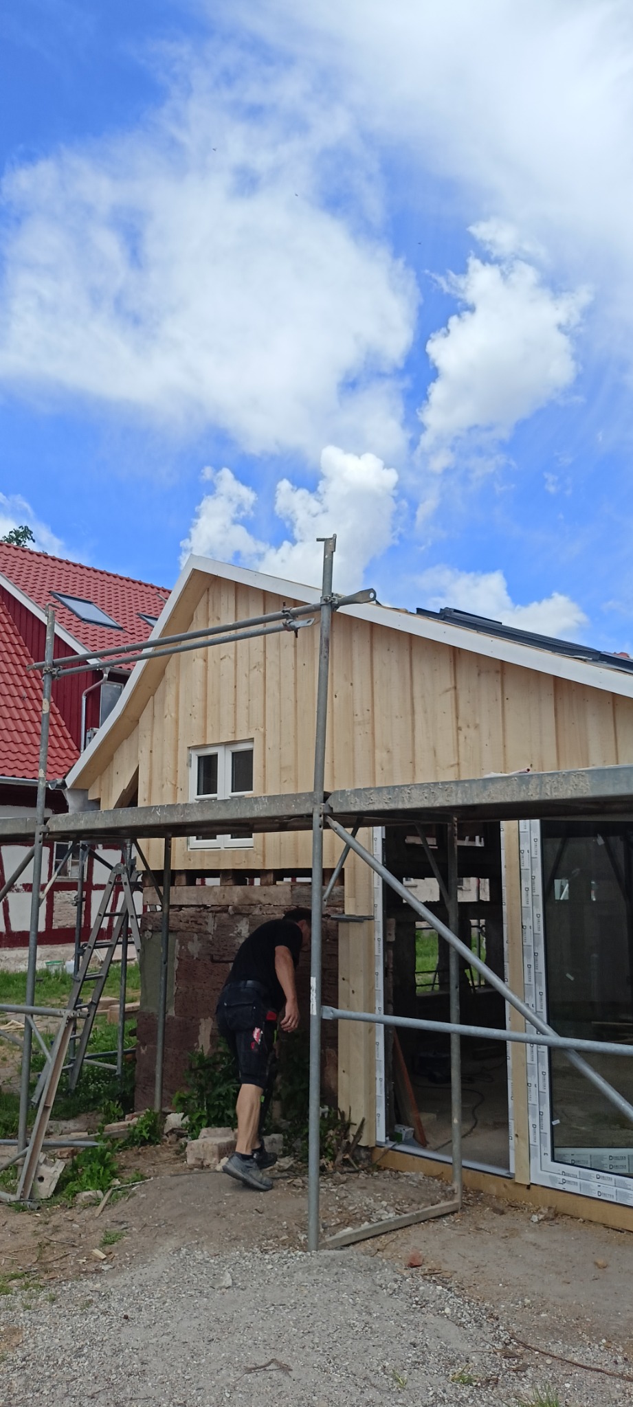 Pracownik na rusztowaniu montuje drewnianą elewację budynku z widoczną konstrukcją okienną i fragmentem sąsiedniego domu z czerwoną dachówką na tle błękitnego nieba z chmurami.