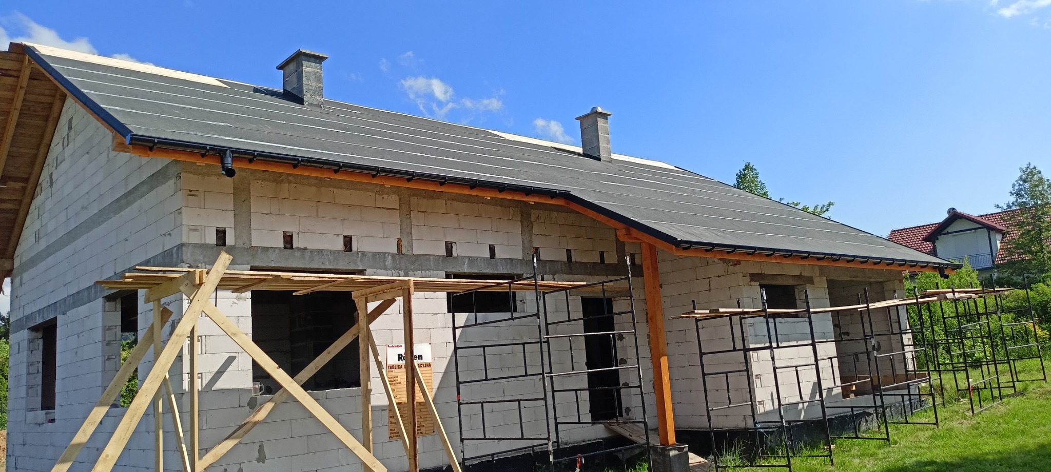 Budynek w trakcie budowy z widocznym dachem pokrytym papą, rusztowaniami i pustakami ceramicznymi, na tle błękitnego nieba.