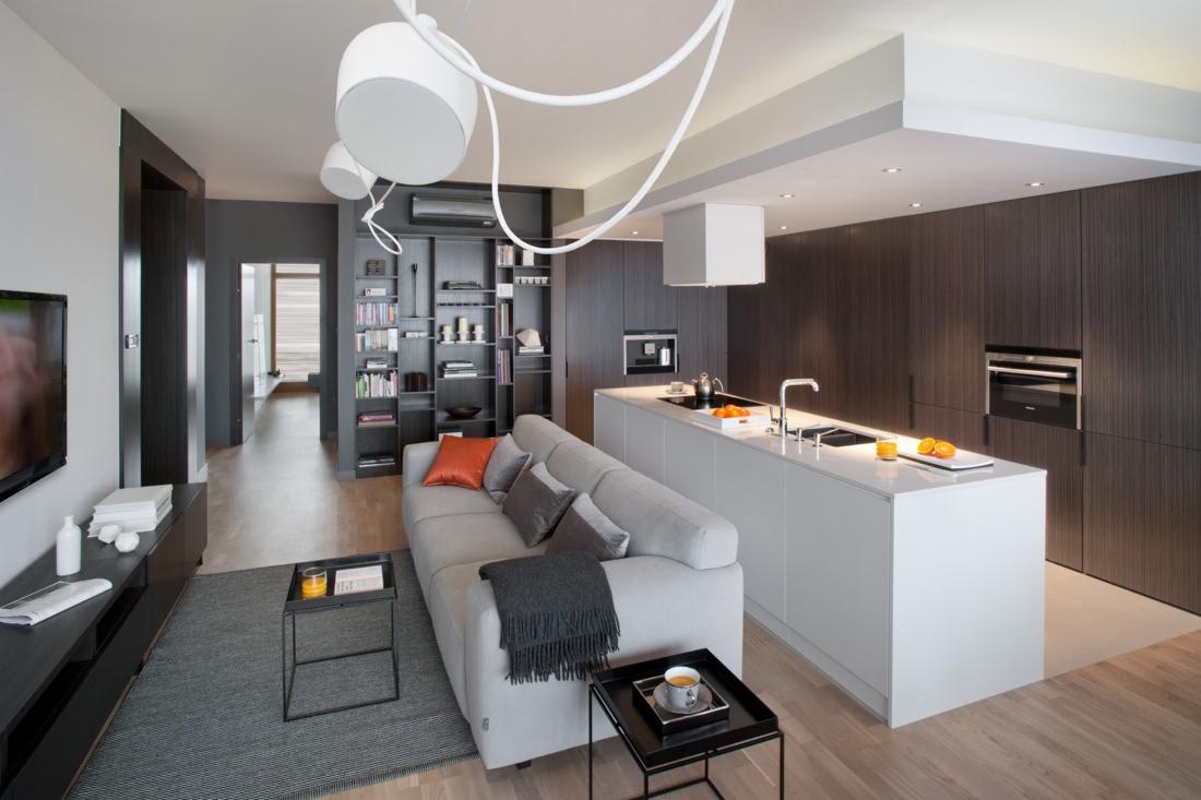 Nowoczesne wnętrze: salon z aneksem kuchennym, szare ściany, drewniana podłoga, biała wyspa kuchenna, szara sofa z pomarańczową poduszką, minimalistyczne oświetlenie.