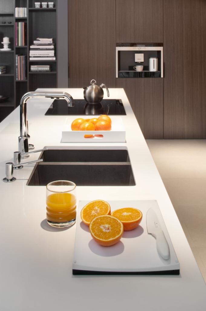 Nowoczesna kuchnia z białym blatem, czarnym zlewem i płytą indukcyjną. Na blacie szklanka soku pomarańczowego, pokrojone pomarańcze na desce i czajnik. W tle zabudowa z ciemnego drewna z wbudowanym...