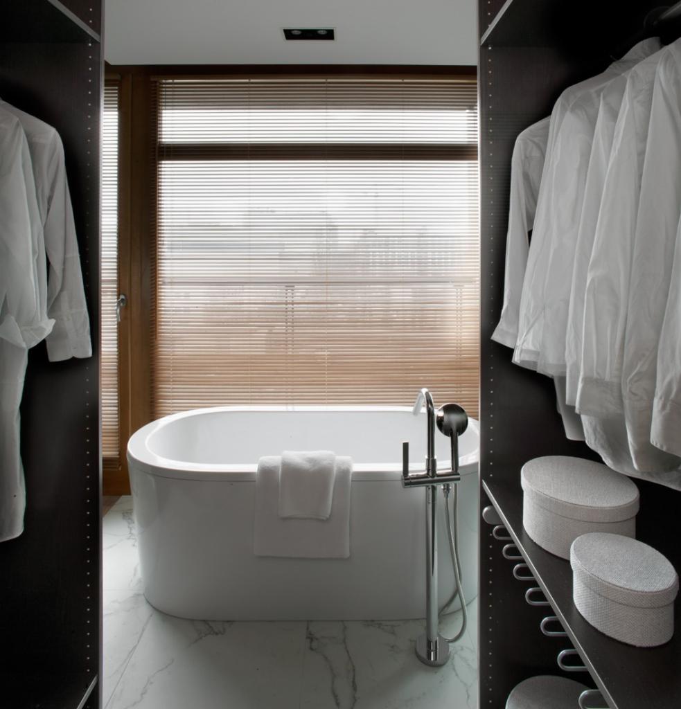 Nowoczesna łazienka z wolnostojącą białą wanną, marmurową podłogą i garderobą z białymi koszulami oraz pudełkami, widok na okno z żaluzjami.