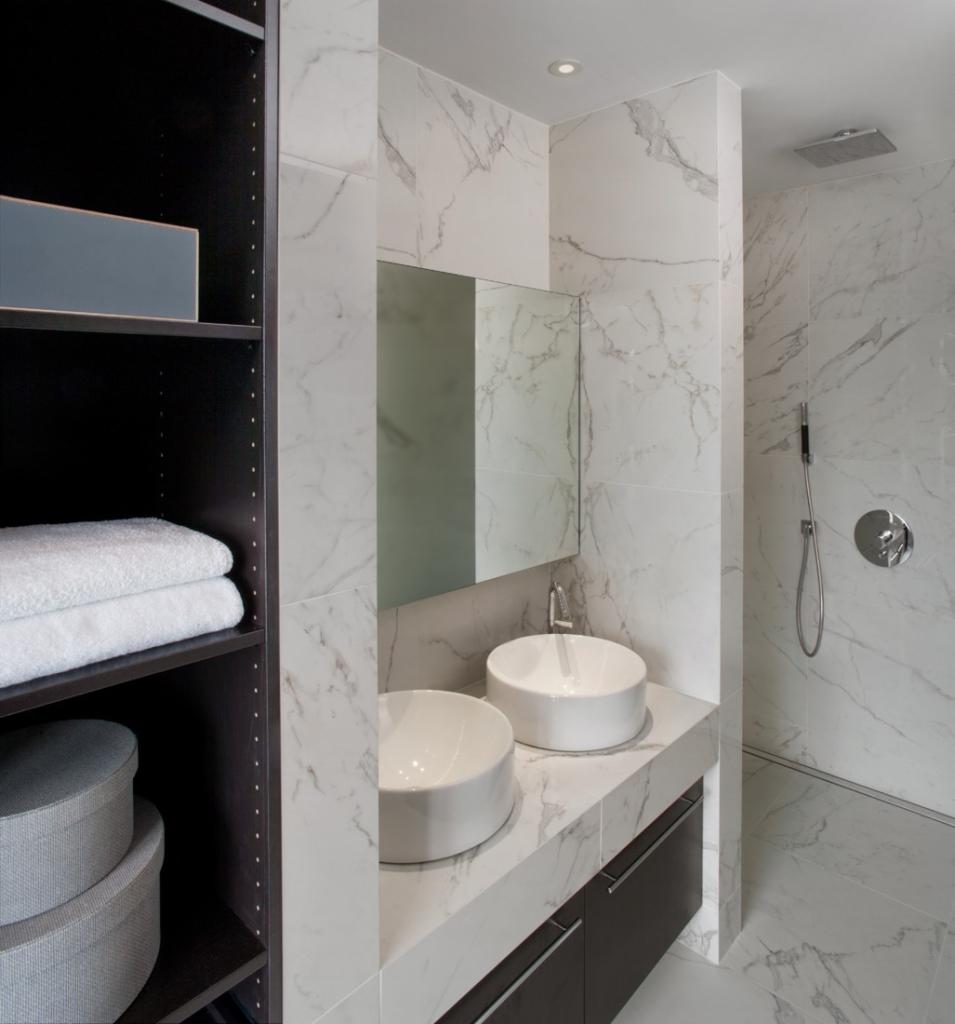 Nowoczesna łazienka z marmurowymi płytkami, dwiema okrągłymi umywalkami nablatowymi, szafką pod umywalką w kolorze grafitowym, lustrem bez ramy i kabiną prysznicową typu walk-in z deszczownicą.