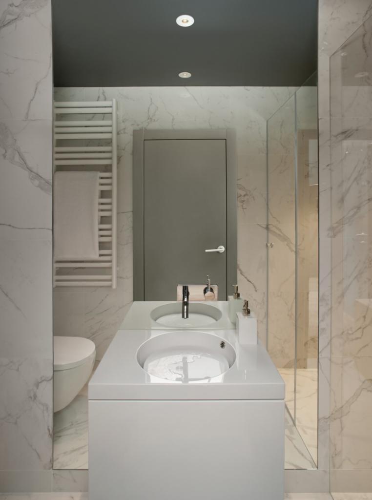 Nowoczesna łazienka w Warszawie z marmurowymi płytkami, grzejnikiem drabinkowym z ręcznikiem, umywalką wpuszczaną w blat i szklanym panelem prysznicowym.