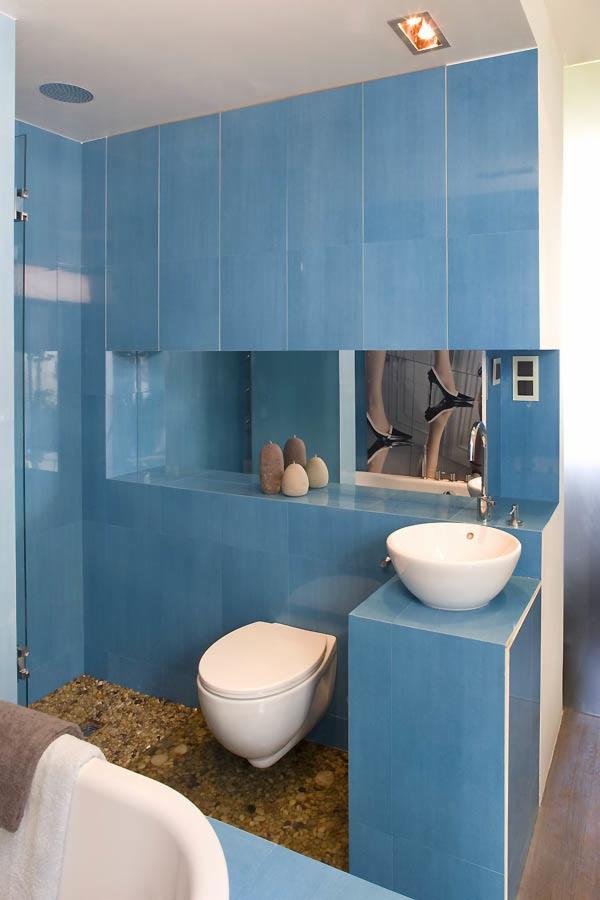 Nowoczesna łazienka z niebieskimi płytkami, okrągłą umywalką nablatową i toaletą wiszącą na tle lustra z odbiciem nóg w szpilkach, podłoga wyłożona drobnymi kamieniami.