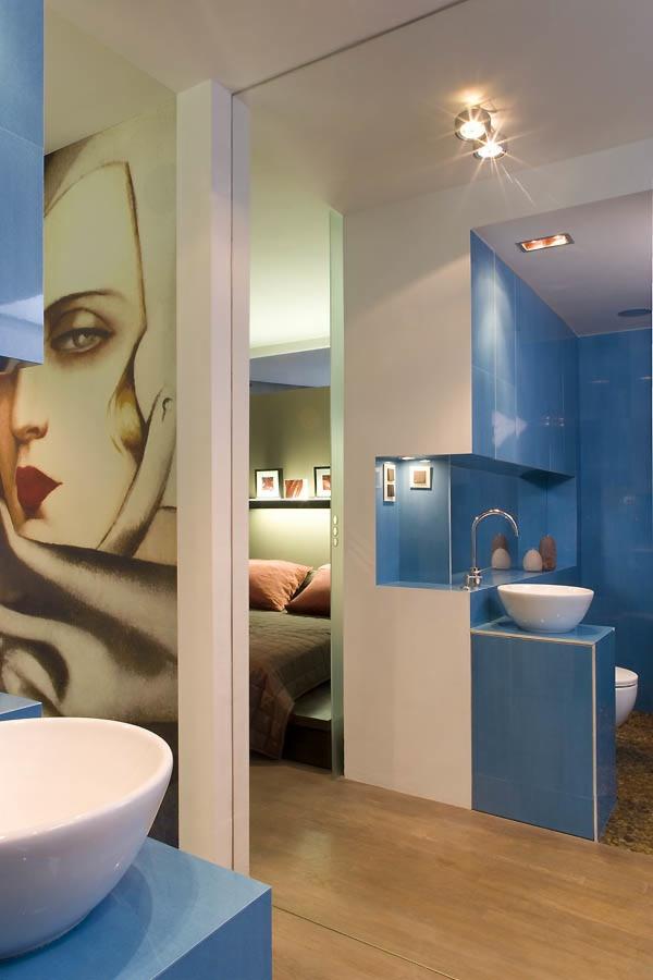 Niebieska łazienka z umywalką nablatową, kafelkami w odcieniach błękitu i szarą ścianą z portretem kobiety, widok na sypialnię z brązową pościelą w tle.