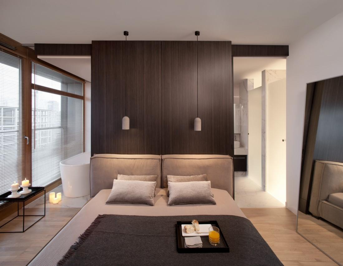 Nowoczesna sypialnia z łazienką en-suite, widok na wannę wolnostojącą, minimalistyczny wystrój z drewnianymi panelami ściennymi i subtelnym oświetleniem, śniadanie na tacy na łóżku, okno z żaluzjami.