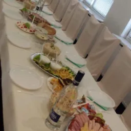 Długi stół z białym obrusem, zastawiony różnorodnymi potrawami, wędlinami, sałatkami i napojami, przygotowany na przyjęcie, z krzesłami w tle i oknem z żaluzjami.