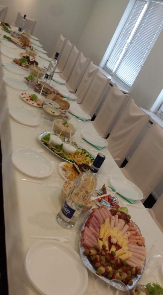 Długi stół z białym obrusem, zastawiony różnorodnymi potrawami, wędlinami, sałatkami i napojami, przygotowany na przyjęcie, z krzesłami w tle i oknem z żaluzjami.