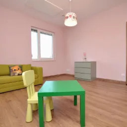 Pokój dziecięcy z różowymi ścianami, drewnianą podłogą, jasnozieloną sofą z poduszką z motywem kota, zielonym stolikiem, żółtym krzesłem, szarą komodą i drewnianymi drzwiami z przeszkleniami.