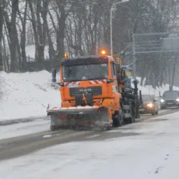Pomarańczowa ciężar&oacute;wka z pługiem odśnieżającym, marki MAN, jedzie po ośnieżonej drodze miejskiej podczas opad&oacute;w śniegu, widoczne inne samochody w oddali.