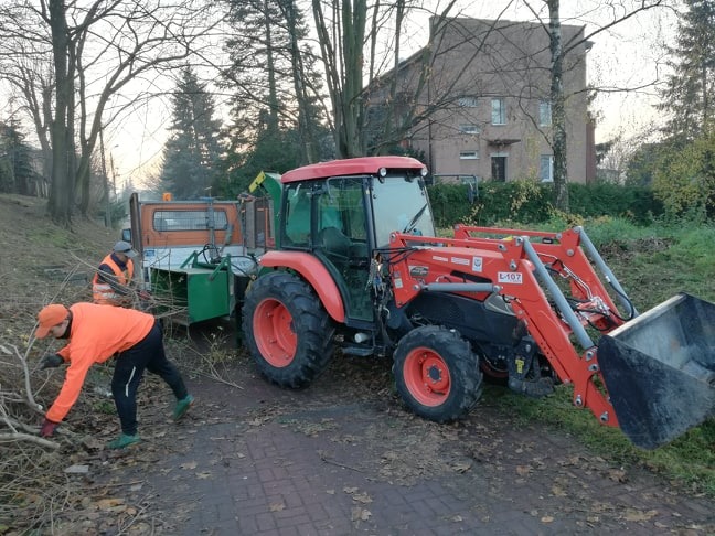 Pomarańczowy traktor Kubota z ładowaczem czołowym i przyczepą z rozdrabniaczem gałęzi, pracownicy w odblaskowych ubraniach zbierają gałęzie po wycince drzew wzdłuż drogi, w tle budynek mieszkalny.