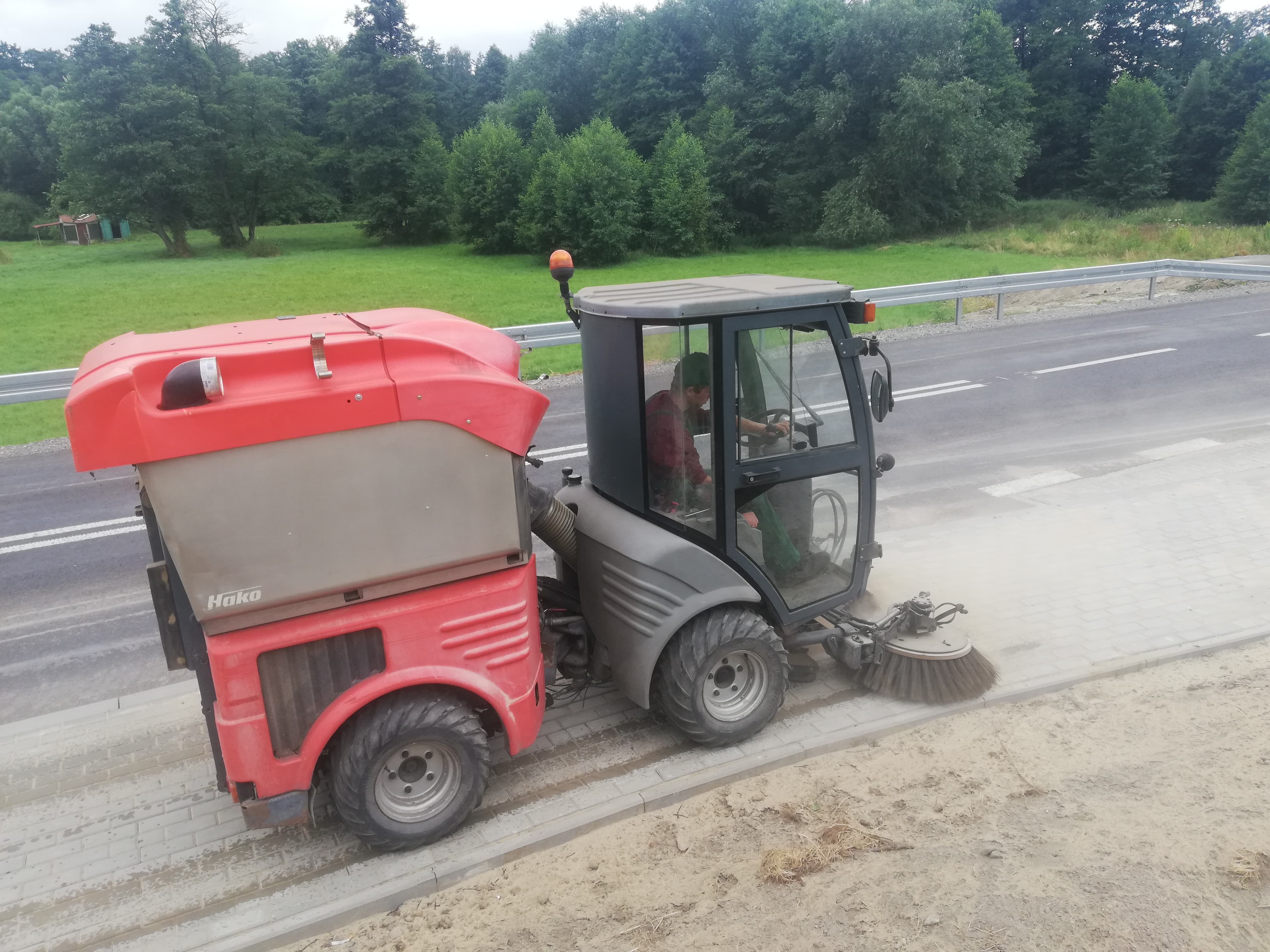 Maszyna czyszcząca Hako zamiata chodnik przy drodze, widoczny operator w kabinie i obracająca się szczotka. W tle drzewa i trawa.