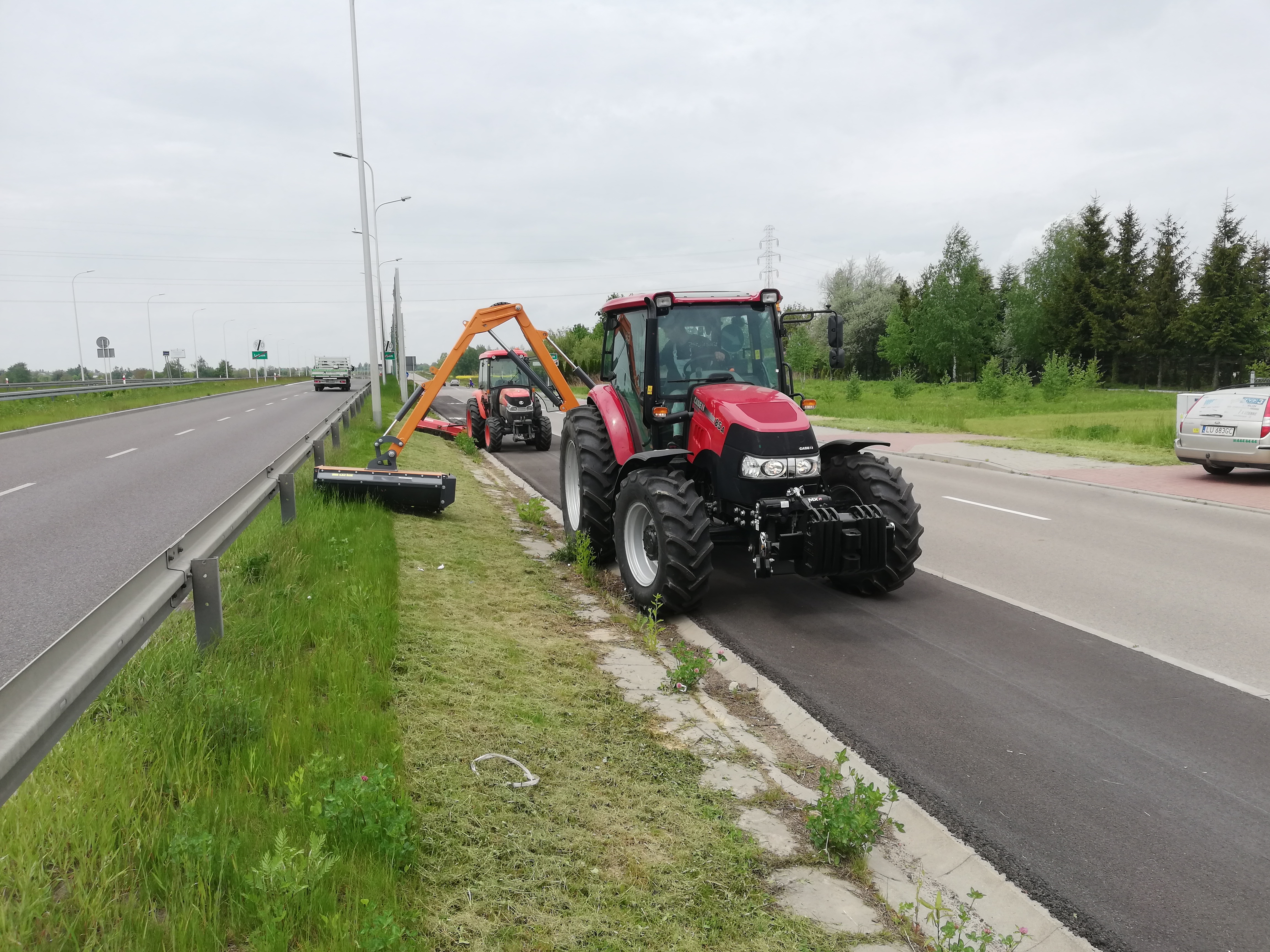 Dwa czerwone traktory z kosiarkami na poboczu drogi ekspresowej, jeden w trakcie pracy, skoszona trawa widoczna obok, w tle ruch samochodowy.