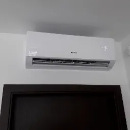 Biały klimatyzator Gree zamontowany na ścianie nad ciemnymi drzwiami w pomieszczeniu.