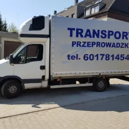 Biała furgonetka z plandeką, na kt&oacute;rej widnieje napis 'TRANSPORT' i numer telefonu 601781454, zaparkowana na ulicy przed budynkiem.