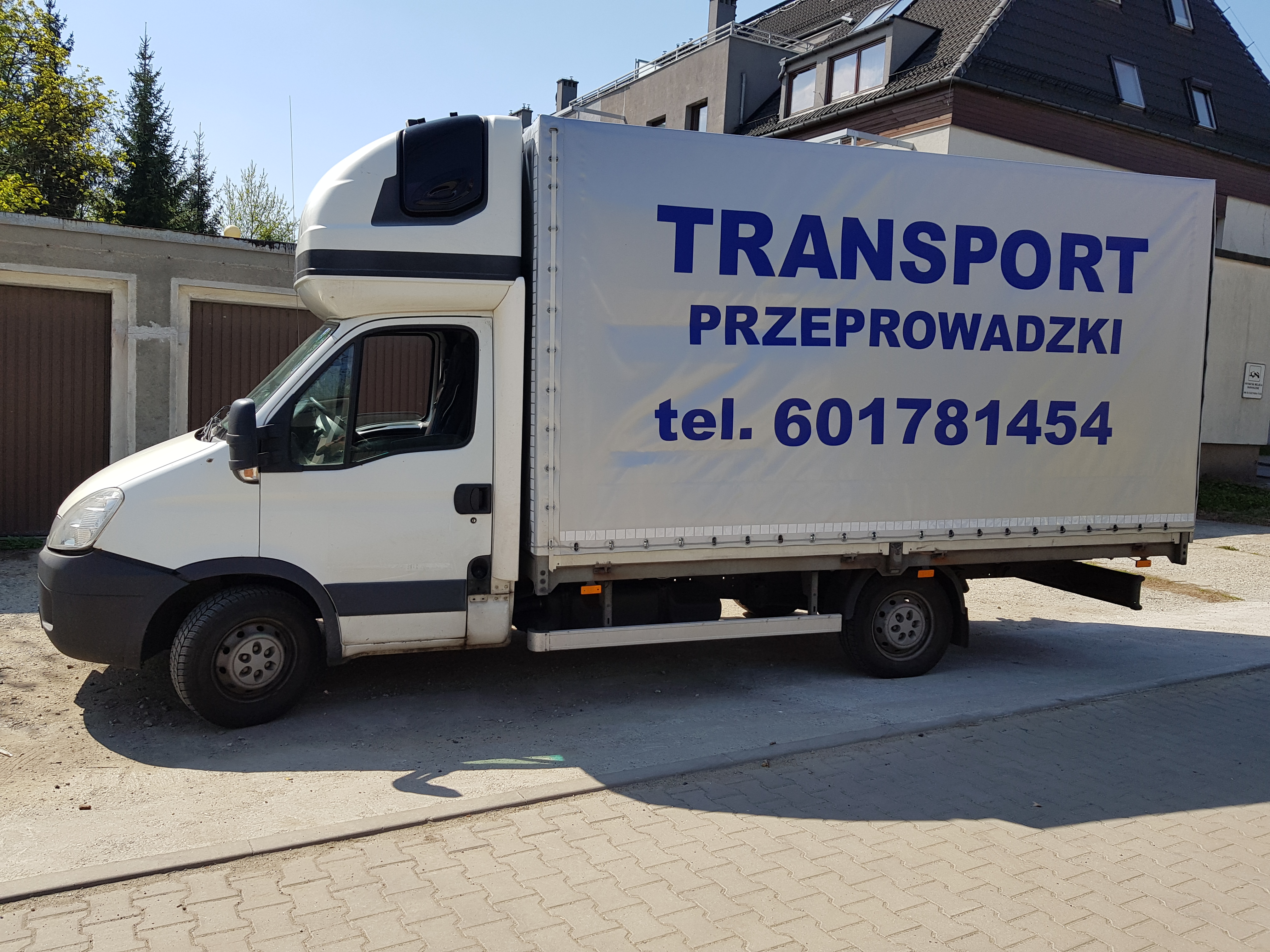 Biała furgonetka z plandeką, na której widnieje napis 'TRANSPORT' i numer telefonu 601781454, zaparkowana na ulicy przed budynkiem.